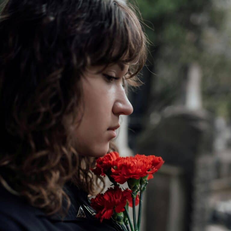A woman in mourning, holding a red rose in front of her face. Neurodivergent people mourn especially hard and it can be a confusing process for us.