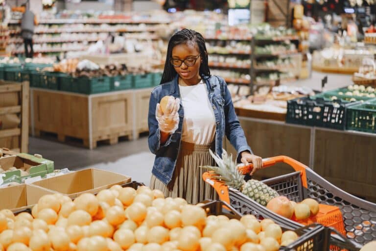 A woman shopping for fresh produce in a grocery store, holding a piece of fruit while pushing a cart filled with fruit.