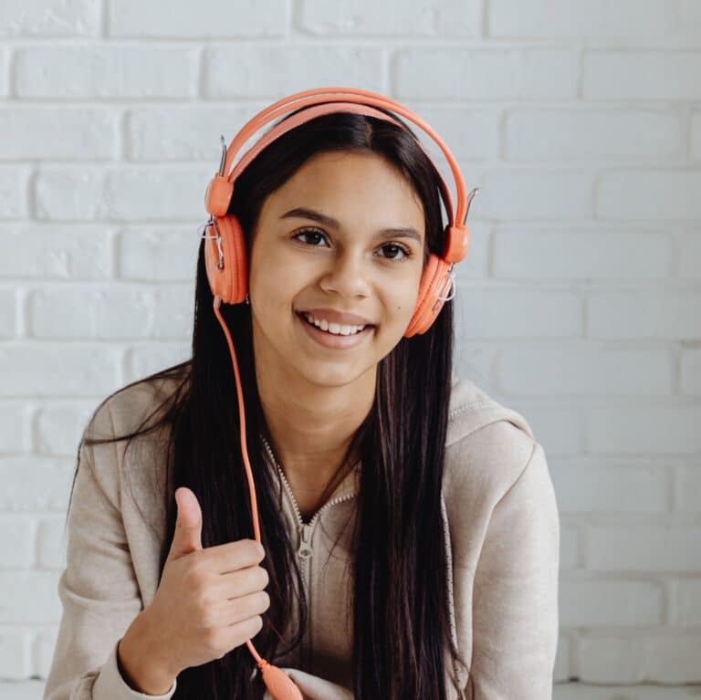 give me alt text A smiling young woman wearing orange headphones gives a thumbs-up in front of a white brick wall.